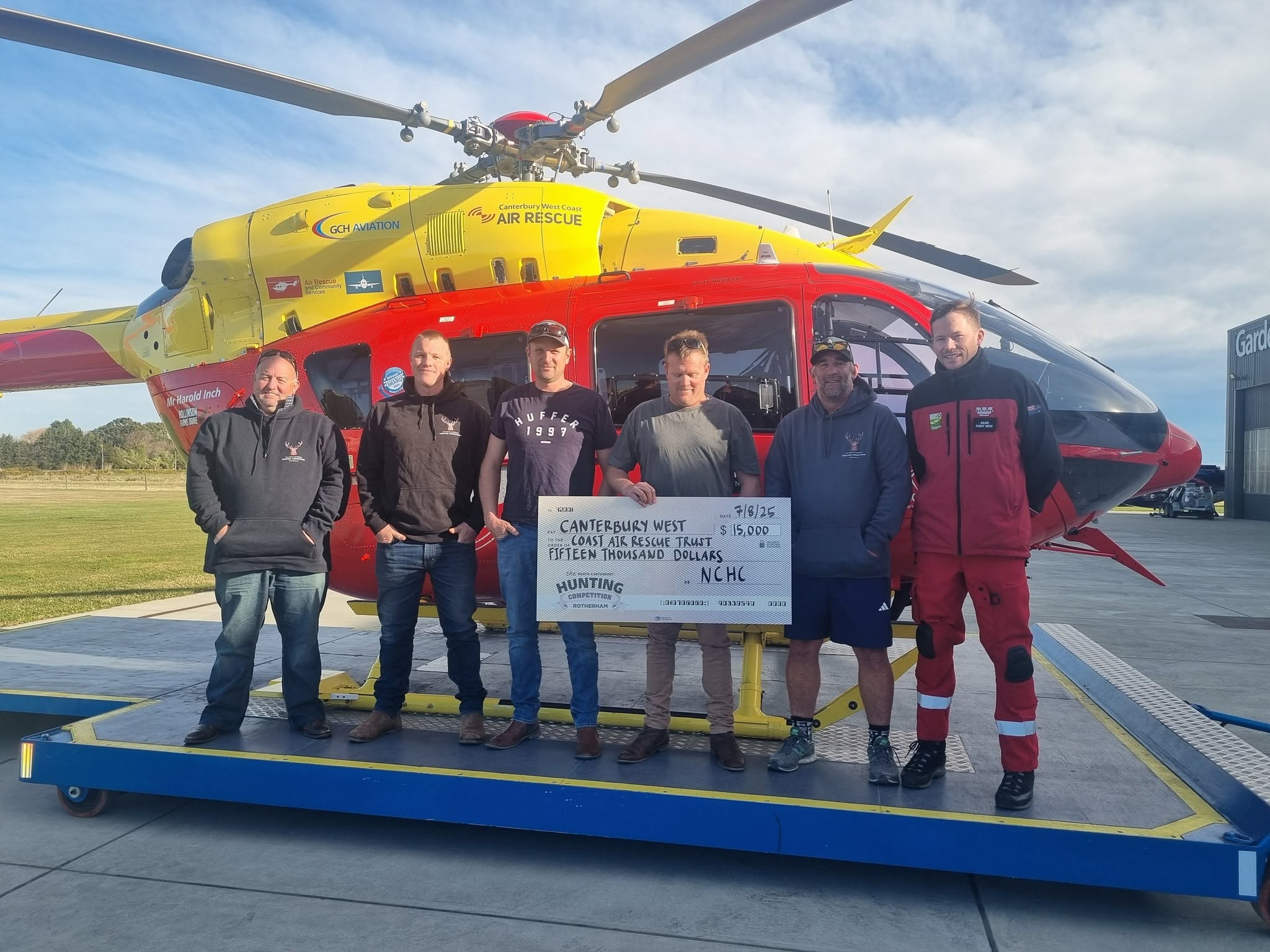 Six men stand in front of a rescue helicopter, holding a large check for $15,000 made out to Canterbury West Coast Air Rescue Trust.