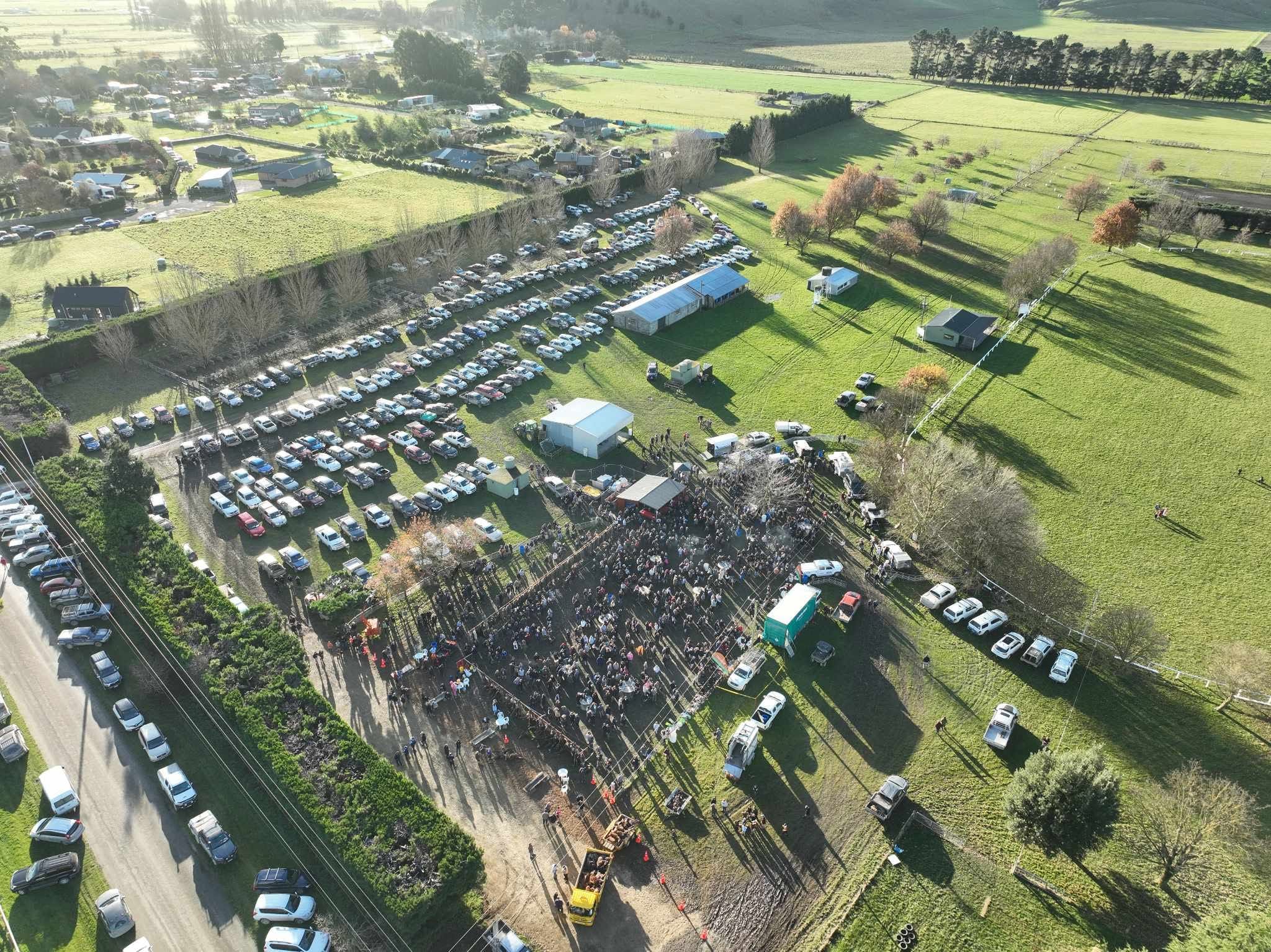 Aerial view of a large outdoor gathering with many parked cars, tents, and people congregating in a fenced area on a grassy field.