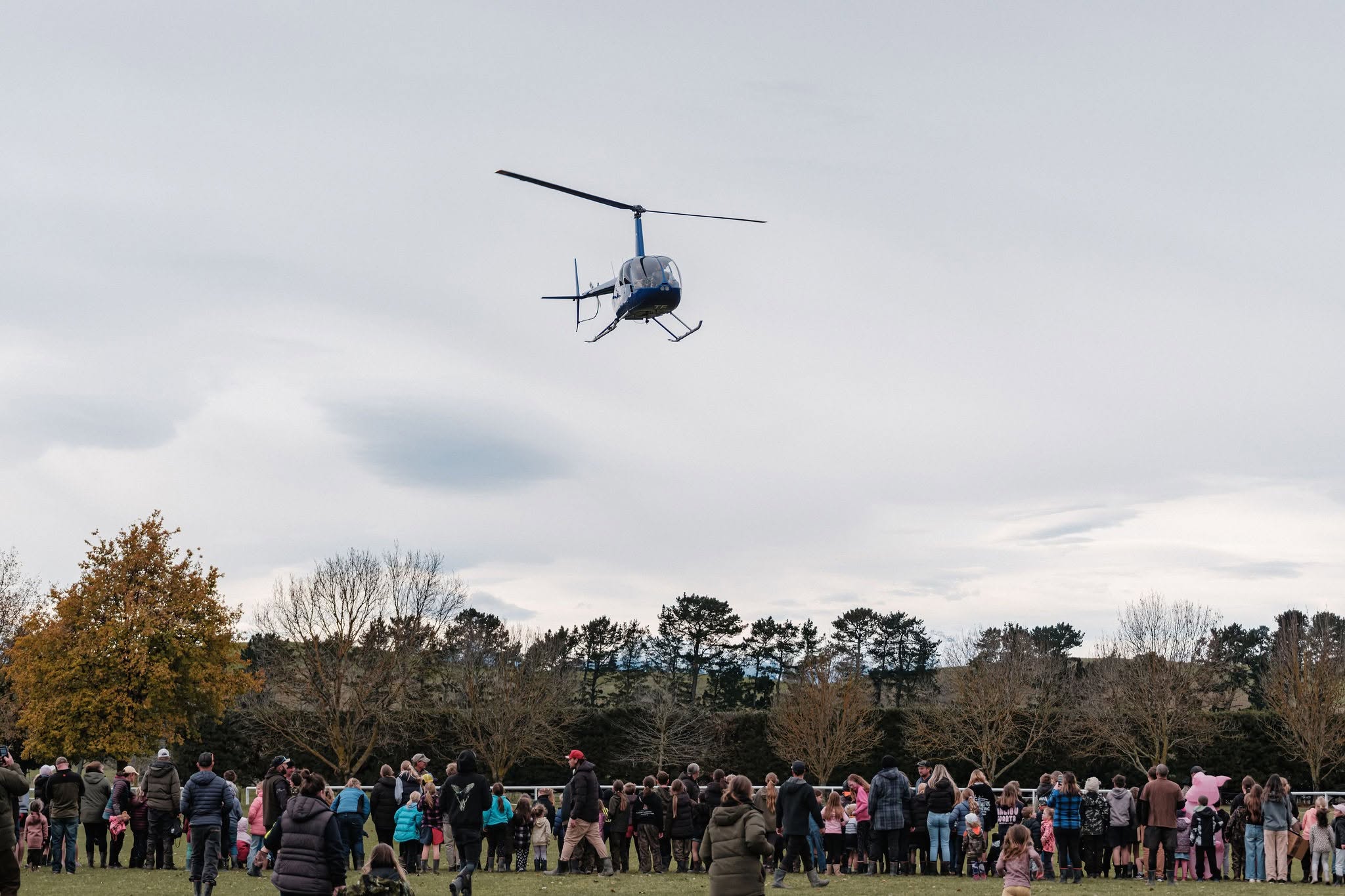 A helicopter hovers above a grassy field while a large crowd of people watch from below on a cloudy day.