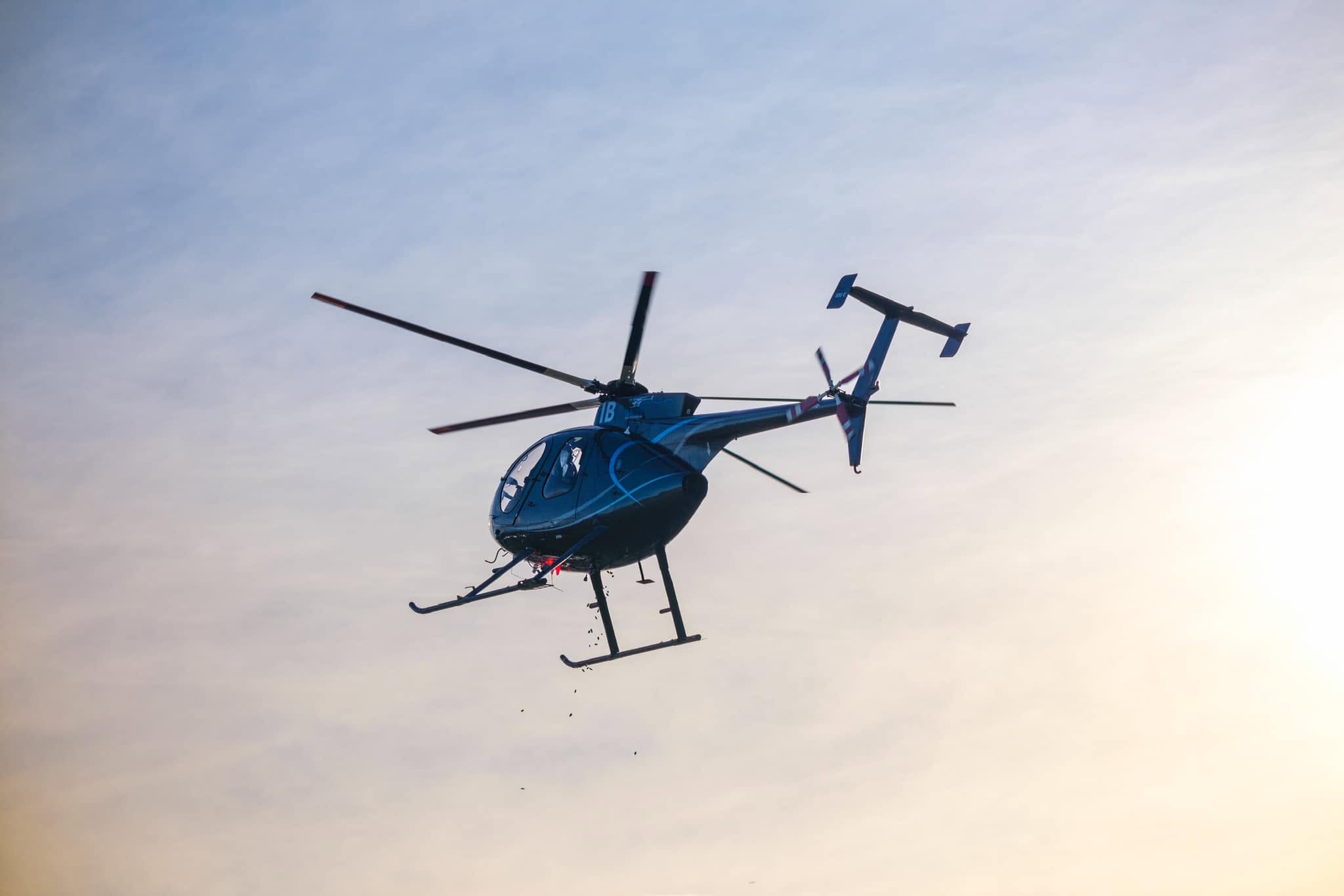A black helicopter flying in the sky during daylight, viewed from below with its rotors in motion against a light blue and yellow sky.