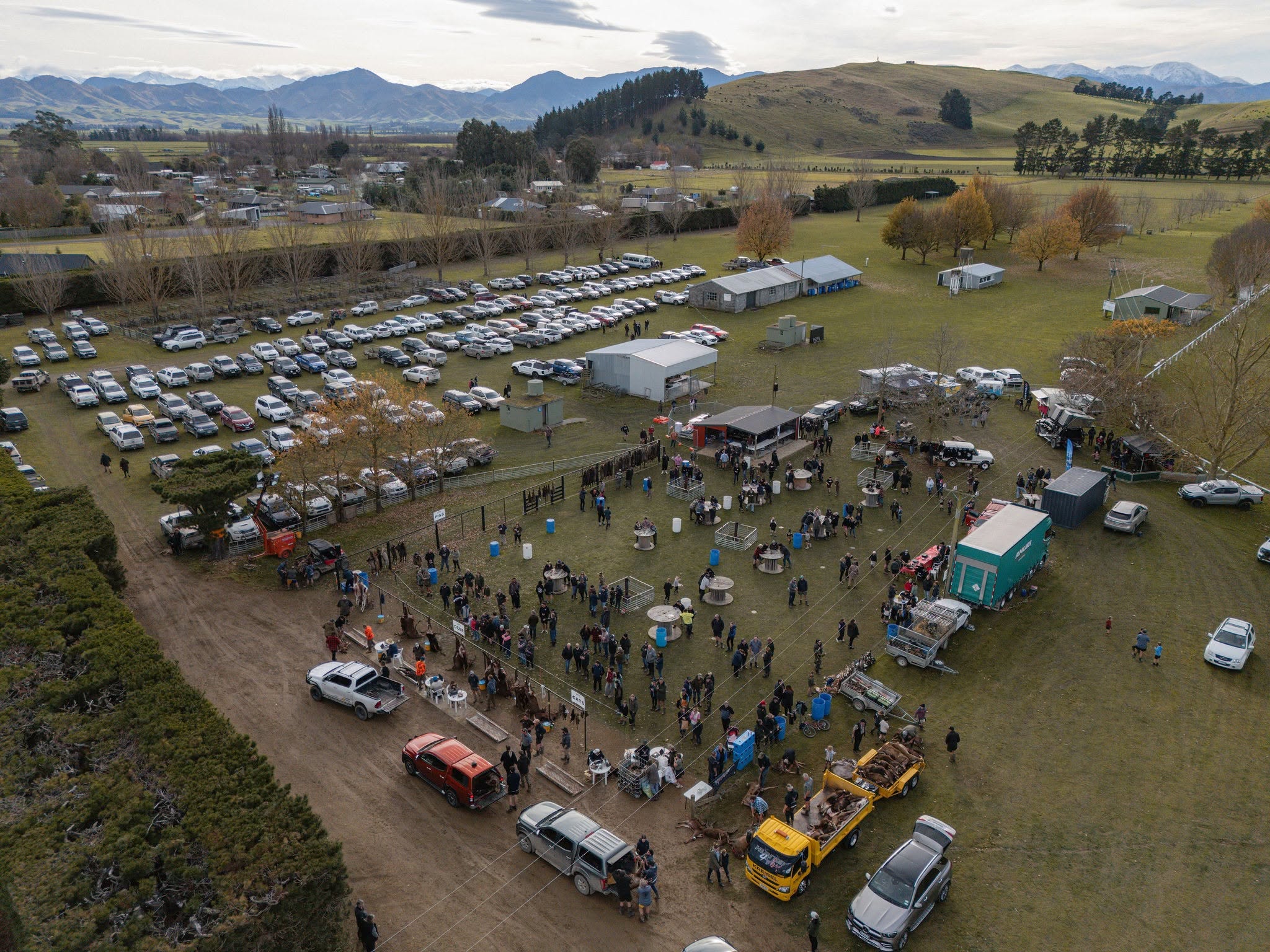 Aerial view of an outdoor event with many people gathered around food trucks and stalls, surrounded by parked cars on a grassy field with buildings and hills in the background.