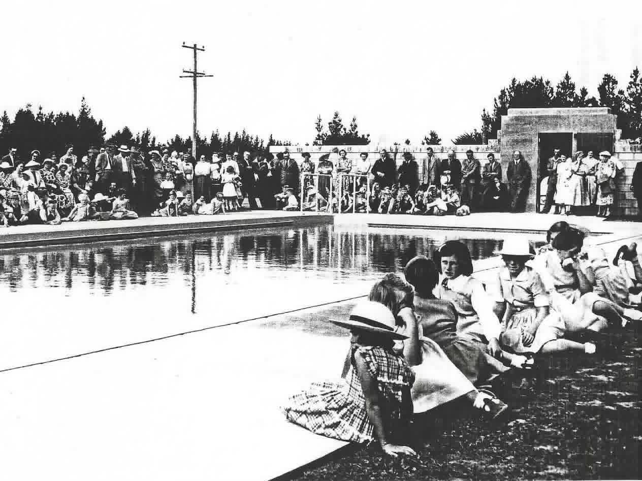A crowd of people, including children and adults, gather around the edge of an outdoor swimming pool during a daytime event.